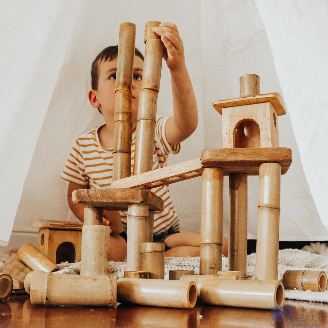 Q Toys Bamboo Building Set, child stacking bamboo lengths into a tower with small wooden houses