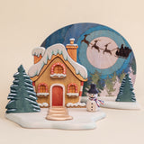 Wooden Christmas scene with a snow-covered gabled house, a moon panel behind it, and small snow trees · view 1