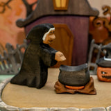 Wooden hooded witch leaning over a black cauldron that sits on a carved wooden fire base · view 1