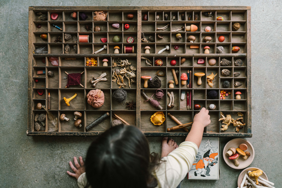 A child reaching into a printer&rsquo;s tray of loose parts: carved mushrooms, acorns, spools, shells, and forest treasures.